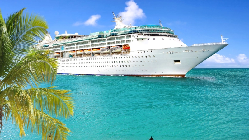 Large cruise ship sailing through clear turquoise tropical waters beside palm trees on a sunny day