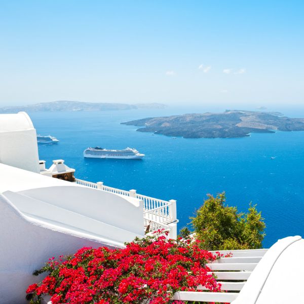 Cruise ships anchored in the blue waters of Santorini's caldera, viewed from a whitewashed hillside with colorful bougainvillea