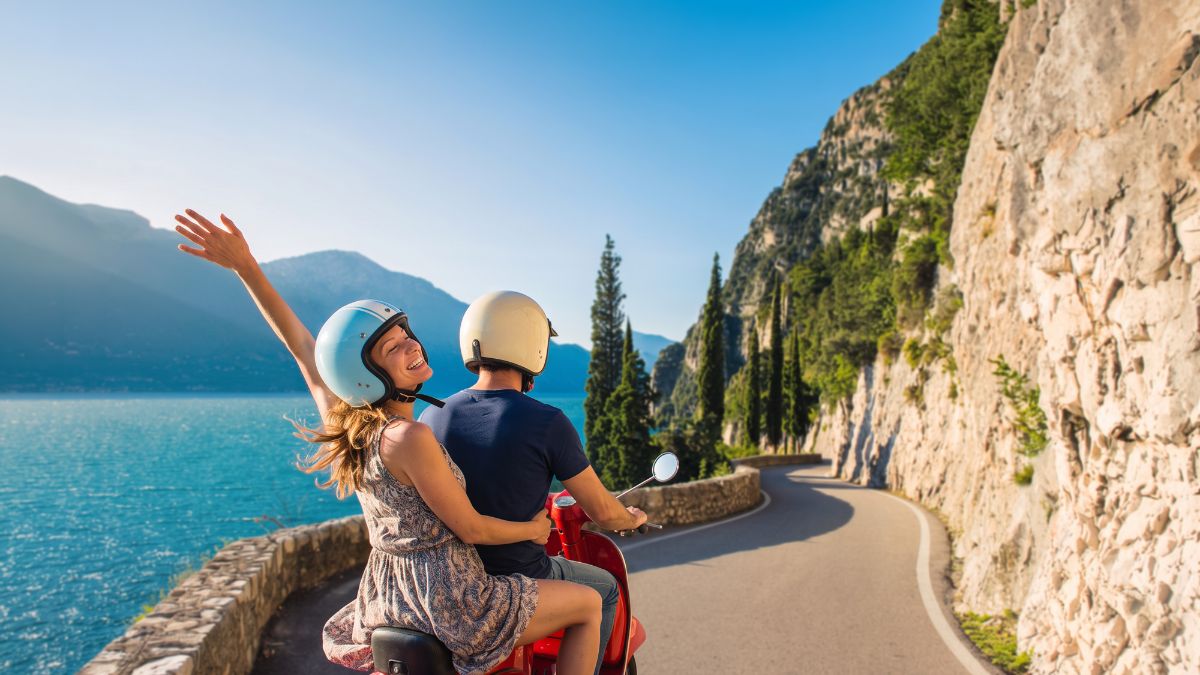 Two people riding a red scooter on a scenic road along a large body of water surrounded by mountains