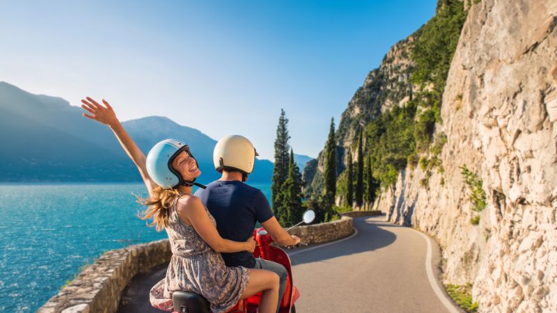 Two people riding a red scooter on a scenic road along a large body of water surrounded by mountains