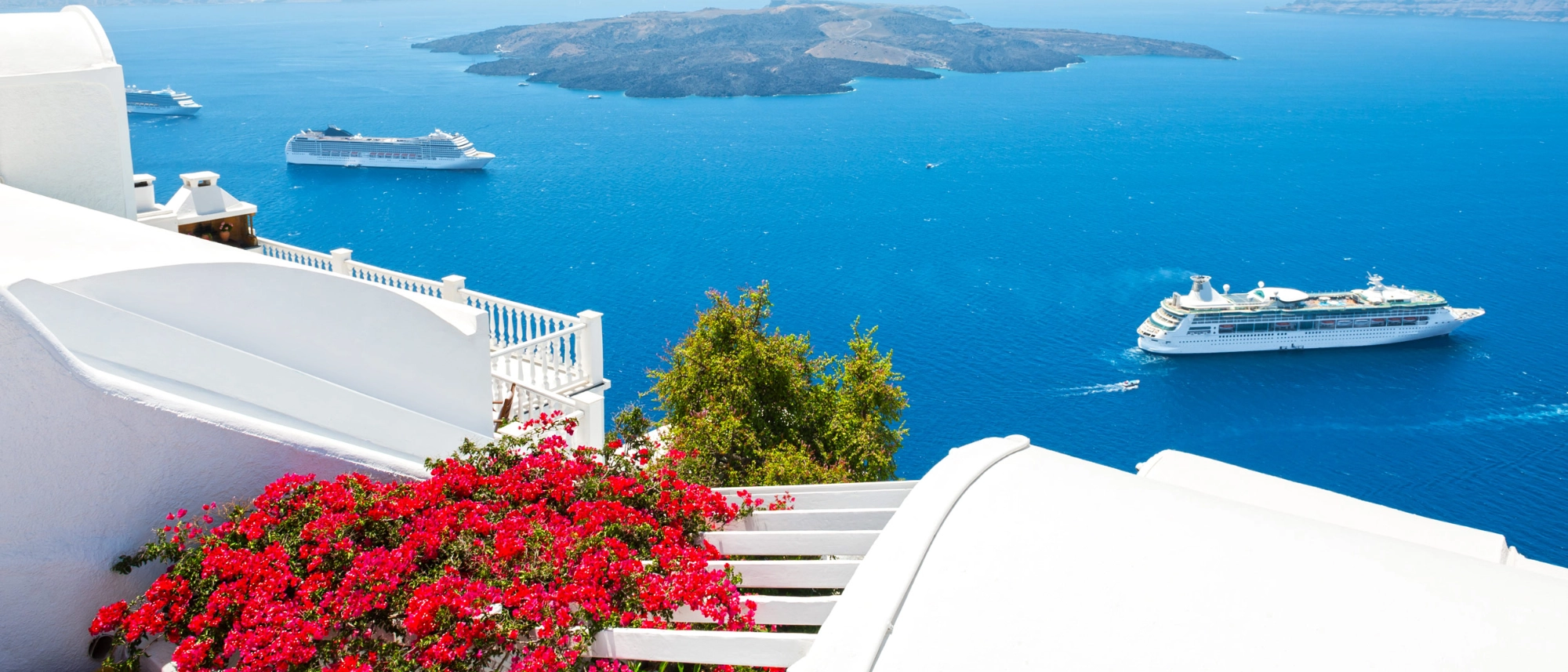 Cruise ships anchored in the blue waters of Santorini's caldera, viewed from a whitewashed hillside with colorful bougainvillea