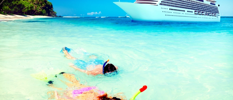 People snorkeling in clear turquoise water near a tropical beach with a cruise ship offshore